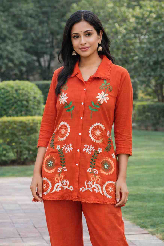 Woman wearing an orange embroidered outfit standing outdoors with greenery in the background
